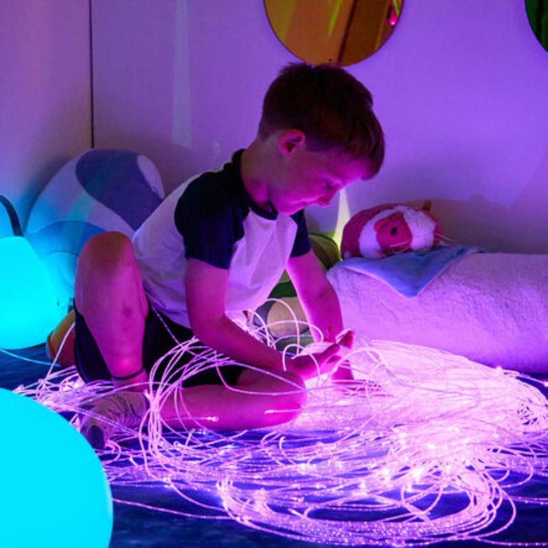 Boy sitting on a starry rug exploring a handful of pink-lit TTS Fibre Optic Tails up close, safe-to-touch strands for calm tactile and visual play