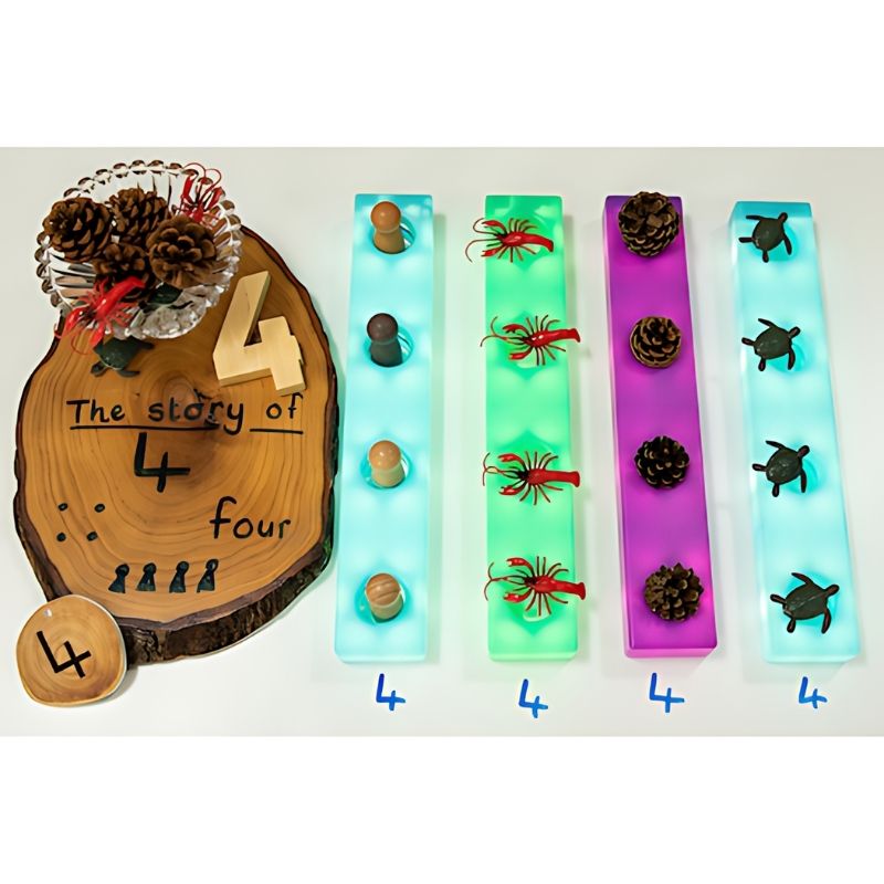 TTS Stack and Build Glow Blocks used as illuminated trays in a loose parts and early maths activity, showing four blocks in blue, green, pink and teal glowing alongside natural objects