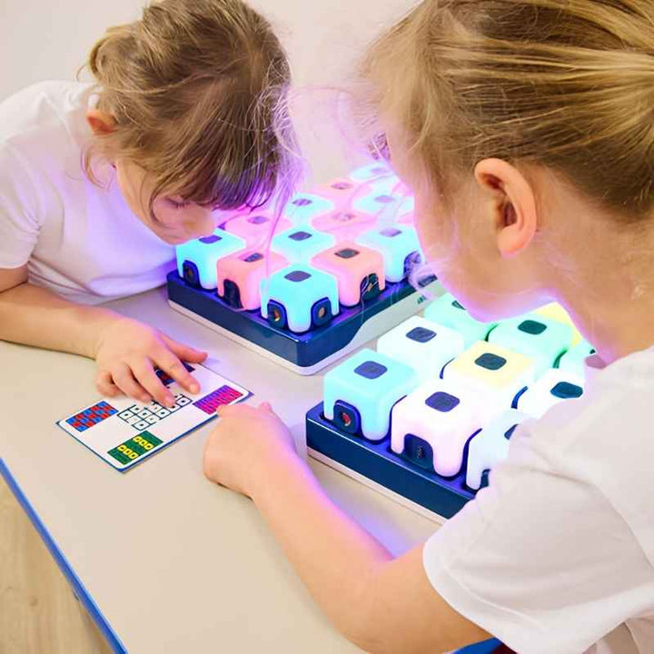 Two children using TTS Glow Sequencing Cubes on charging docks at a classroom table, with one child referencing a printed activity card to recreate a colour sequence pattern, showing the structured curriculum learning application