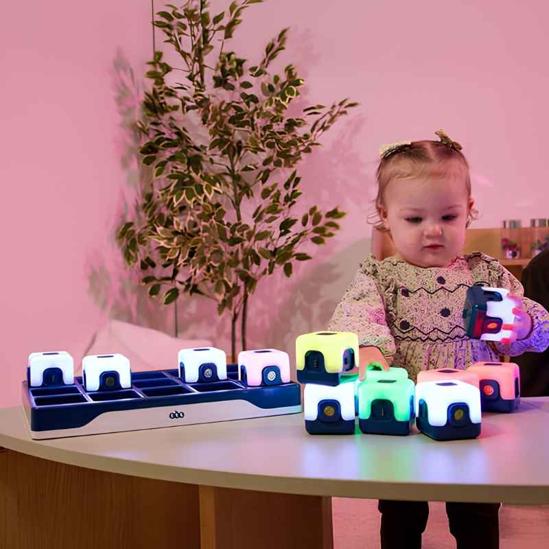 TTS Glow Sequencing Cubes with the included charging dock in the background and a baby exploring individual glowing cubes on a table, showing the rechargeable set in a home or nursery setting