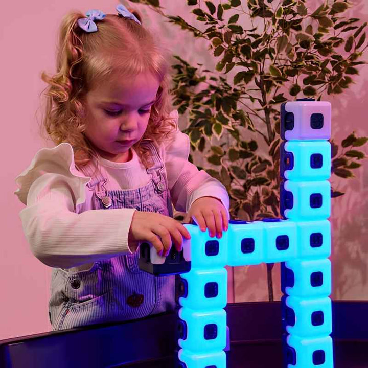 Young child building a glowing blue arch and tower with TTS Glow Sequencing Cubes on a tray table, demonstrating open-ended magnetic construction and early spatial reasoning