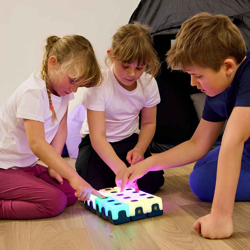 Three school-age children collaborating around a flat grid of TTS Glow Sequencing Cubes in a classroom setting, demonstrating group sensory learning and turn-taking