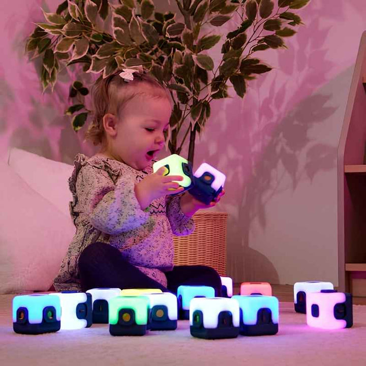 Toddler closely examining a single TTS Glow Sequencing Cube while a full set glows in multiple colours on the floor, supporting early sensory exploration from 10 months