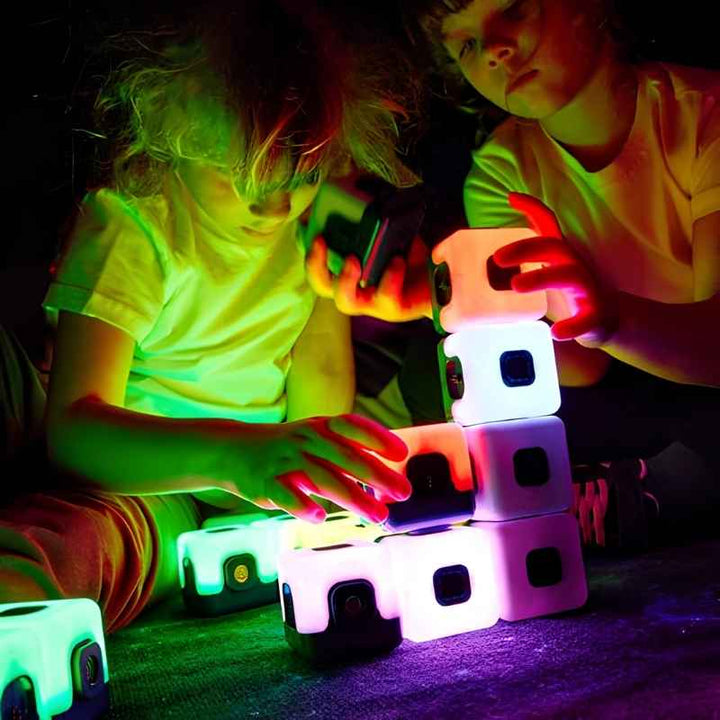 Two children building a tower with TTS Glow Sequencing Cubes in a darkened room, hands lit by the glow of the cubes, demonstrating collaborative sensory light play