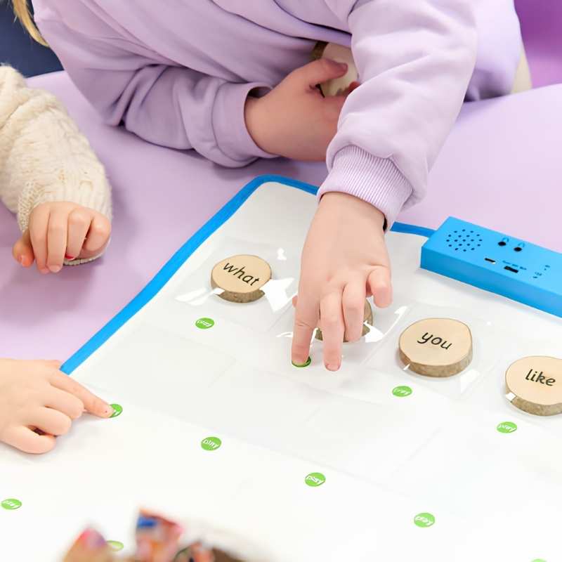 Two young children using the TTS Interactive Talking Mat on a classroom table, placing wooden word tokens into pockets to support speaking and language development