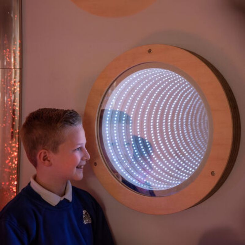 Smiling boy standing beside the TTS Light Up Circular Infinity Mirror glowing with white LED rings, a wall-mounted sensory mirror for visual exploration in sensory rooms