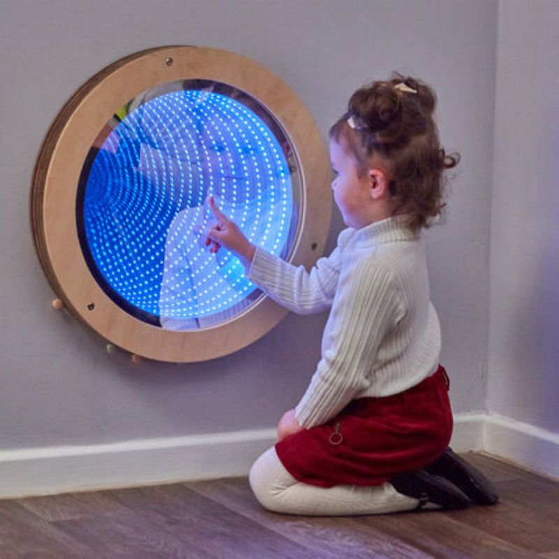 Young girl kneeling to point into the blue-lit TTS Light Up Circular Infinity Mirror, its LED rings creating an endless tunnel illusion in a wall-mounted wooden frame