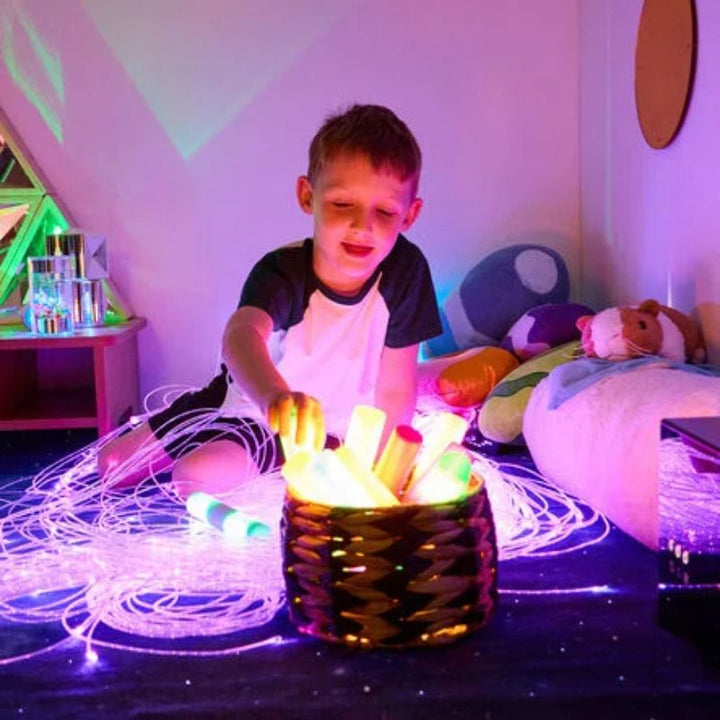 Boy reaching into a basket of glowing TTS Light Up Glow Cylinders on a starry rug surrounded by fibre optic strands, enjoying calm sensory light play
