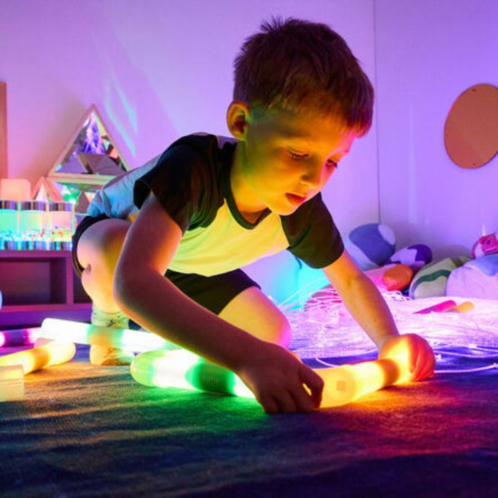 Boy kneeling to line up TTS Light Up Glow Cylinders end to end on the floor, building fine motor skills and creative construction through open-ended play