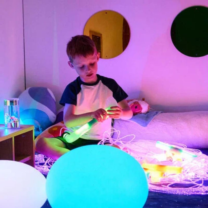 Boy sitting in a sensory corner examining a glowing TTS Light Up Glow Cylinder, with more cylinders, fibre optic strands and glow balls around him