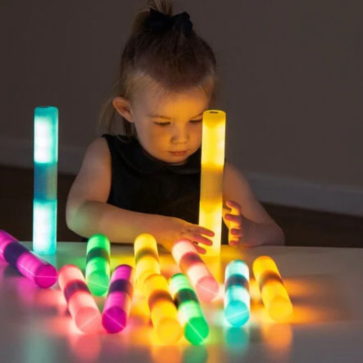 Young girl standing glowing TTS Light Up Glow Cylinders upright on a table beside a colourful row of the full set, exploring colour sorting and stacking