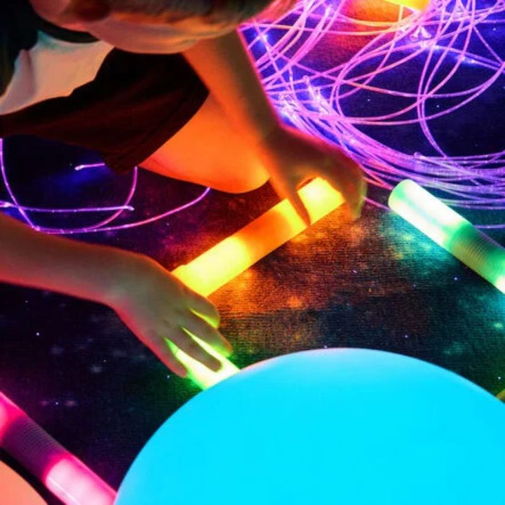 Top-down view of a child’s hands arranging glowing TTS Light Up Glow Cylinders on a starry rug beside a light-up ball and fibre optic strands