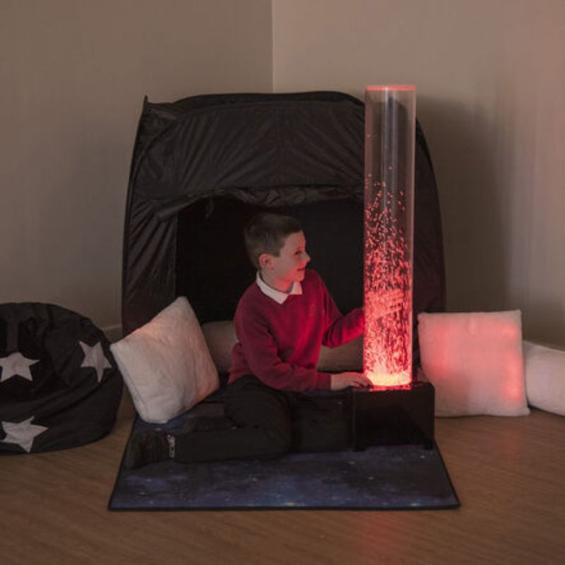 Boy sitting at the entrance of the TTS Mini Pop-Up Sensory Pod beside a glowing red bubble tube, creating a calm corner with cushions for sensory regulation