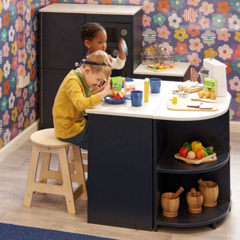 Two children collaborating at the TTS Modern Living Kitchen Island Set in an early years classroom, with one seated on a stool and role play food and accessories spread across the worktop