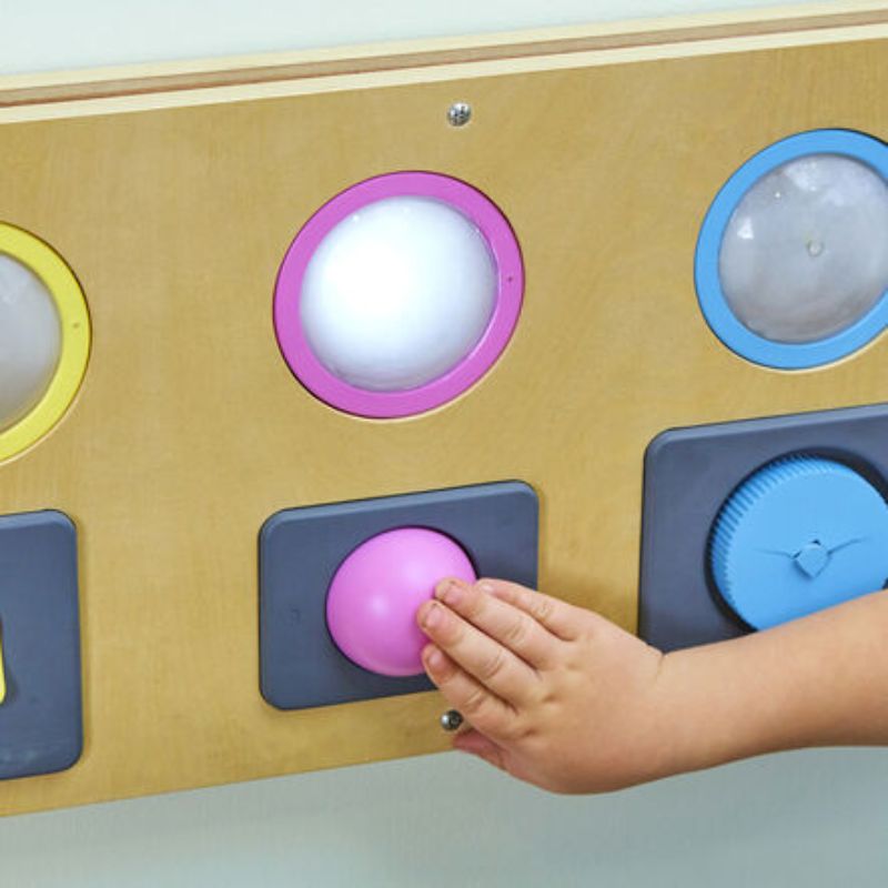 Child pressing the pink push button on the TTS Switch and Press Fine Motor Board as a dome light glows above, an interactive sensory wall panel for cause and effect