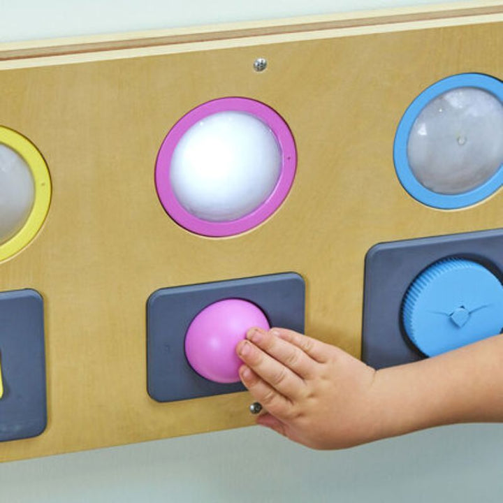 Child pressing the pink push button on the TTS Switch and Press Fine Motor Board as a dome light glows above, an interactive sensory wall panel for cause and effect