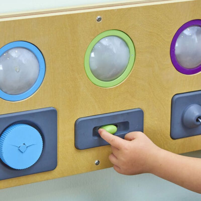Child pushing the green slider on the TTS Switch and Press Fine Motor Board beside a blue turning dial, building finger strength and hand control through hands-on play