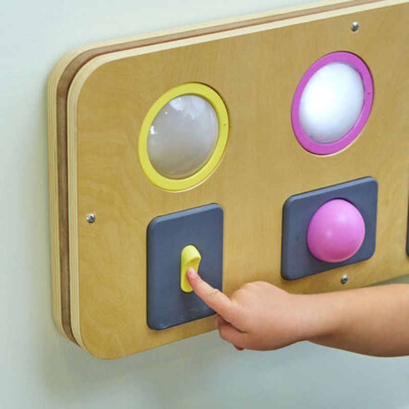 Child flicking the yellow toggle switch on the TTS Switch and Press Fine Motor Board, a wall-mounted cause-and-effect panel with light-up domes for fine motor practice