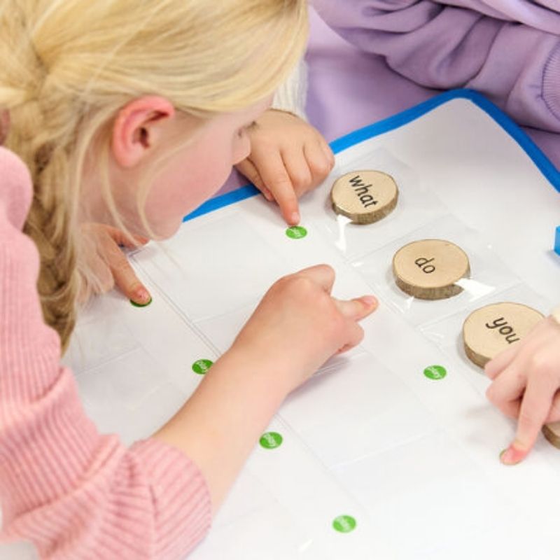 Two children using the TTS Interactive Talking Mat with wooden word tokens to build vocabulary and spoken language skills, pressing the green play buttons on the recordable mat