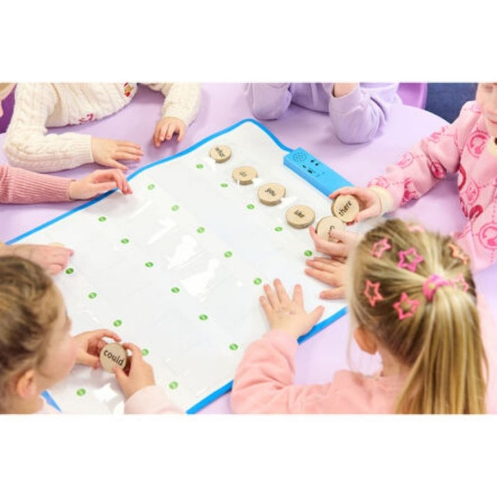Four children seated around the TTS Interactive Talking Mat on the floor, placing wooden word tokens into pockets during a collaborative spoken language and communication session