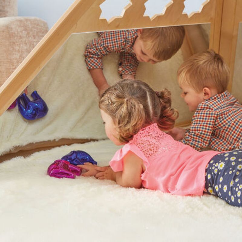 Three toddlers lying on a soft rug inside the TTS Wooden Pyramid Den, exploring their reflections in the mirrored wall during sensory play