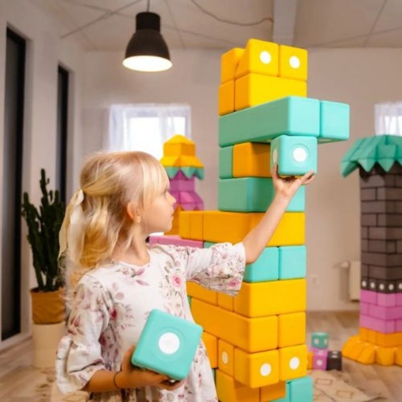 Girl picking up a large 10cm TUKI BIG magnetic foam block from the Builder 110 set, showing the whole-body physical engagement and bilateral coordination that large-format floor building with the 110-piece largest BUILDER magnetic foam set develops