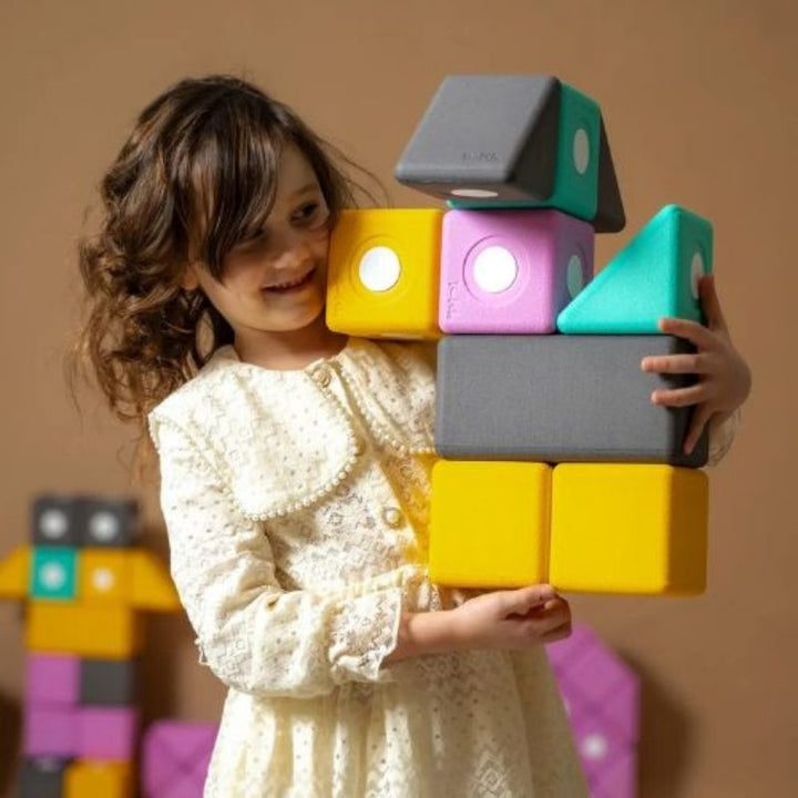 Girl lifting a large 10cm TUKI BIG Smart magnetic foam block from the Smart 10 set, showing the bilateral coordination and proprioceptive engagement that large-format Smart shape foam block construction develops