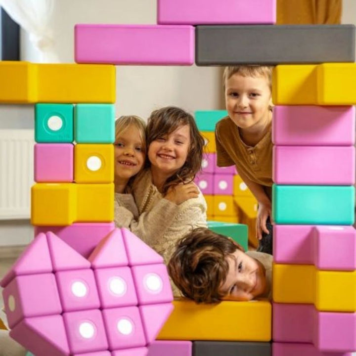Four children playing with the TUKI BIG Smart Magnetic Blocks Smart 30, showing the large-format 10cm SMART shape foam block floor construction the 30-piece pyramid-vocabulary set enables in early years and institutional settings