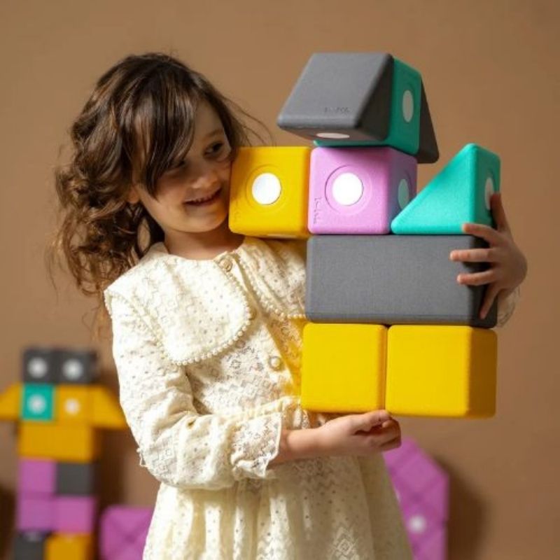 Girl lifting a large 10cm TUKI BIG Smart magnetic foam block from the Smart 75 set, showing the bilateral coordination and proprioceptive engagement that large-format Smart shape floor building with the 75-piece pyramid-vocabulary group set develops