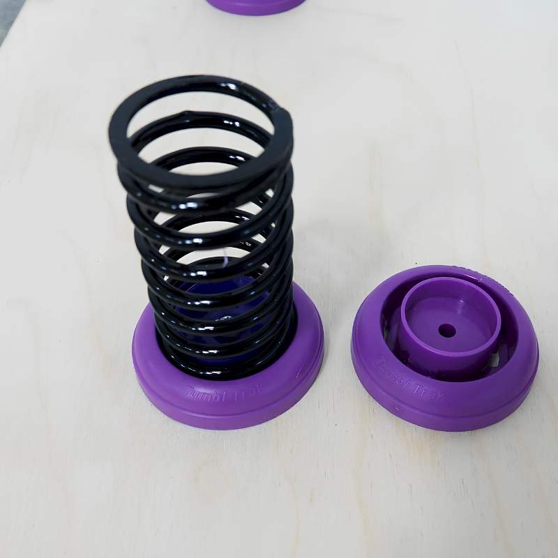 Close-up of a Tumbl Trak gymnastics spring floor kit component showing a black high-carbon steel coil spring seated in a purple plastic retainer bottom cap, with a second purple top cap placed alongside on a light wood panel surface