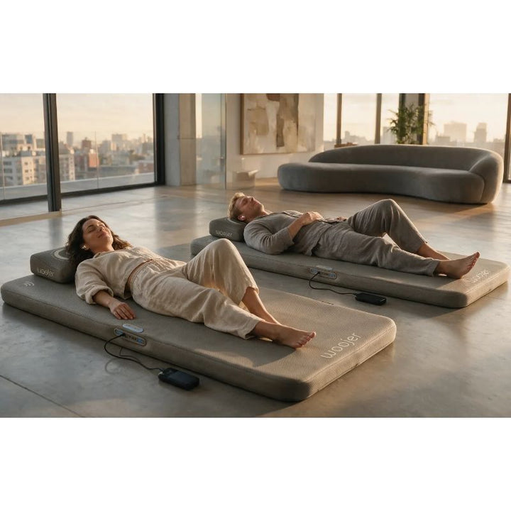 Two people relaxing on Woojer MAT Pro vibroacoustic mats in a bright living space, using the vibrating mat for calm at-home sessions