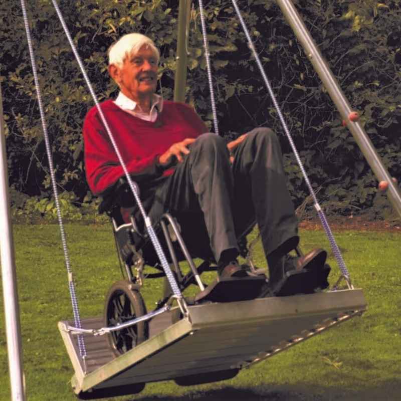 An adult wheelchair user swinging on the TFH Wheelchair Platform Swing with Frame, showing the full accessible swing frame in use