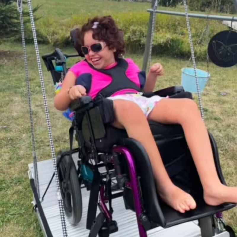A child secured in her wheelchair on the TFH Wheelchair Platform Swing, an accessible sensory swing set up outdoors for all abilities