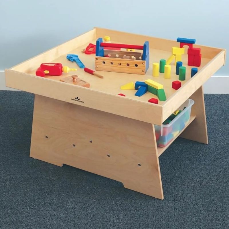 Side view of the Whitney Brothers Big Wide Discovery Table with toys on the surface, showing the raised perimeter lip, wide stable base, and open under-table storage for toddler classrooms