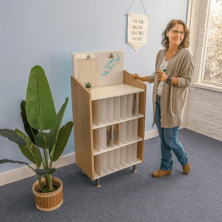 Adult using the Whitney Brothers elevatED Mail and Message Center in a classroom setting, showing the 24 vertical slots, removable dividers, and the dry-erase board with clips at the top