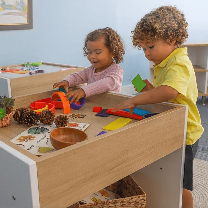 Two toddlers playing together at the Whitney Brothers elevatED Toddler Discovery Table, showing the 26" wide surface that accommodates side-by-side parallel play and the elevatED Collection design