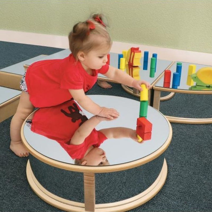 Close-up of an infant exploring the Whitney Brothers Infant Floor Mirror Set, showing the shatterproof acrylic mirror surface of a birch plywood block at floor level