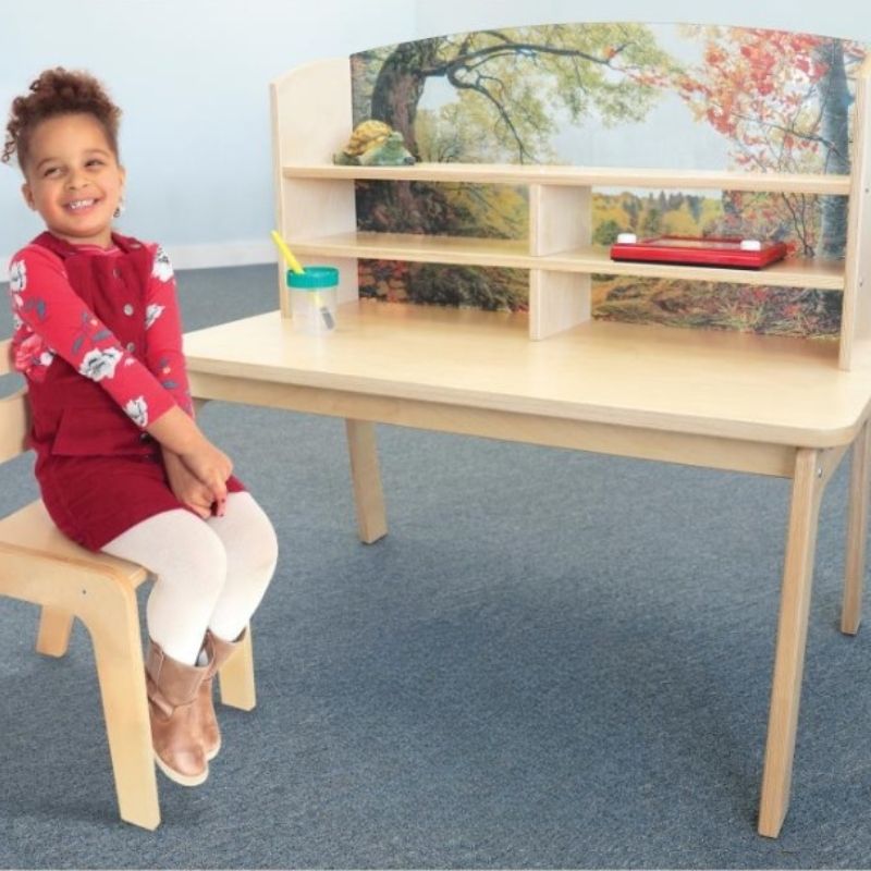 Happy child seated at the Whitney Brothers Nature View Writing Center, showing the forest scene acrylic back panel and the 20" desk surface of the award-winning biophilic writing desk