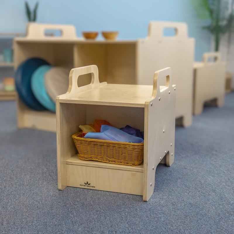 Whitney Brothers Stack-Able Seat in a natural birch finish shown in an early learning classroom, with open storage compartment holding a wicker basket of coloured manipulatives and additional Stack-Able units arranged in the background