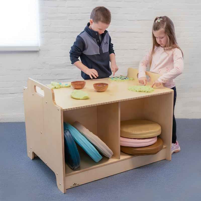 Two children standing at the Whitney Brothers Stack-Able Table engaged in a play dough activity, with floor cushions stored in the divided below-surface compartments