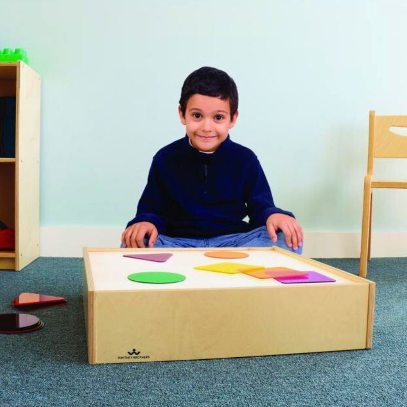 Child sitting and playing with the Whitney Brothers Superbright LED Tabletop Light Box placed on the floor, exploring translucent materials on the illuminated portable light box surface